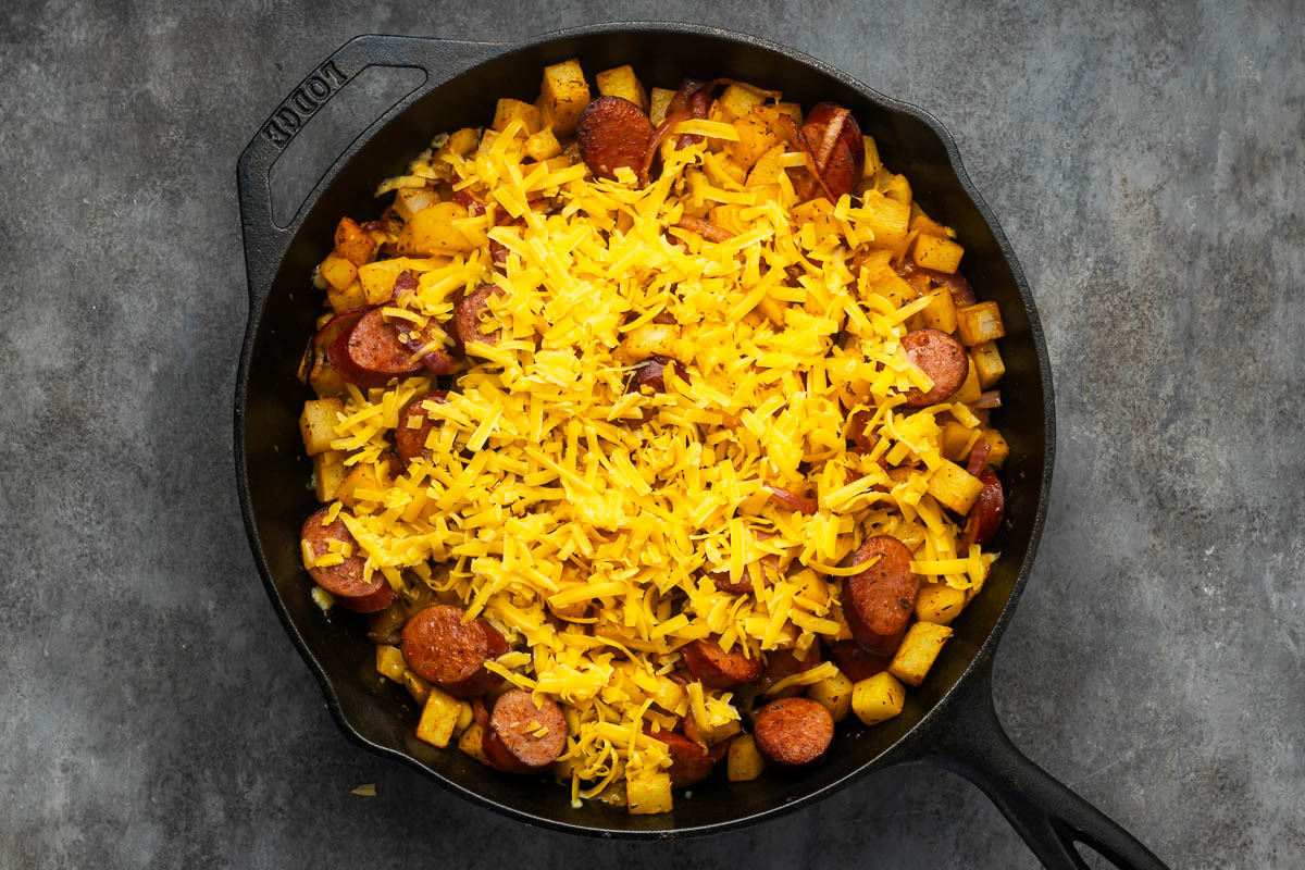 A cast iron skillet filled with sliced sausage, diced potatoes, and topped with a layer of shredded cheddar cheese.