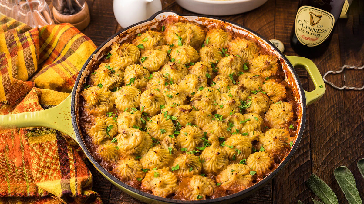 A skillet of shepherds pie topped with piped mashed potatoes and garnished with chopped herbs, placed on a wooden table with a plaid cloth and a bottle of Guinness in the background.