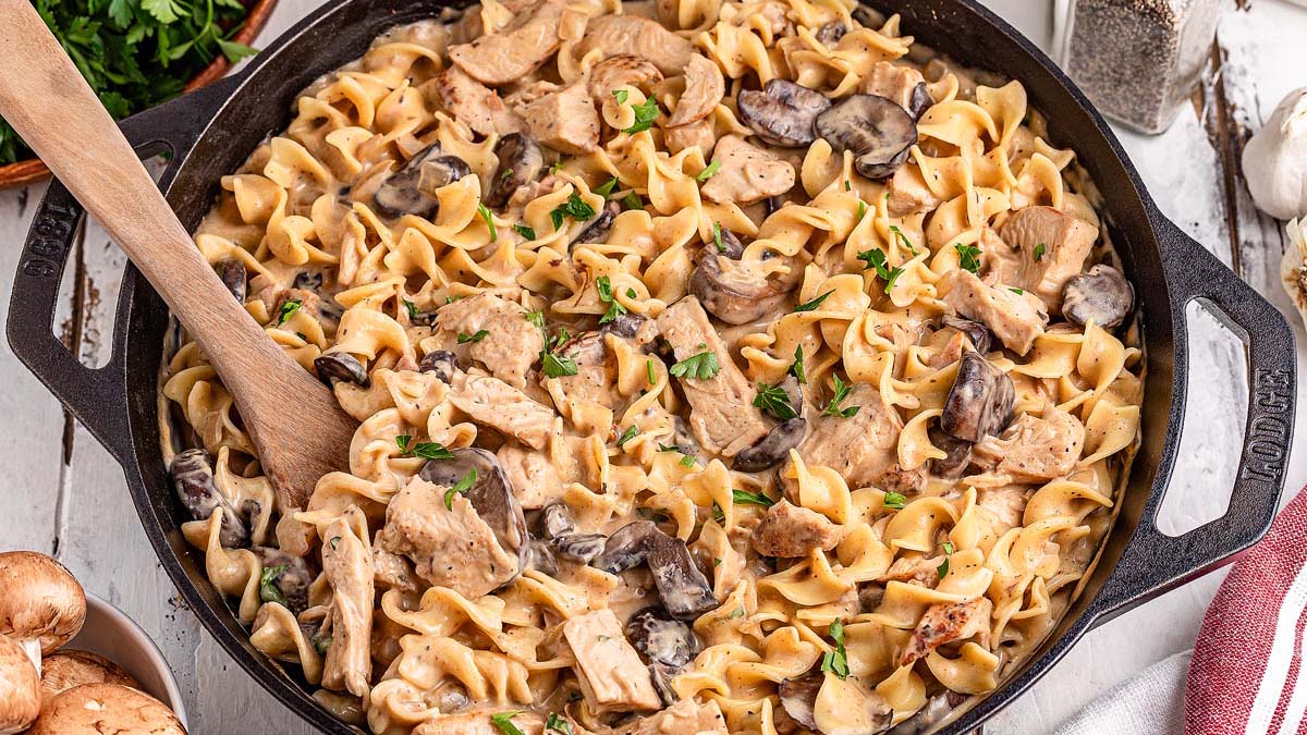 A cast iron skillet filled with creamy chicken and mushroom pasta, garnished with herbs, with a wooden spoon resting inside.