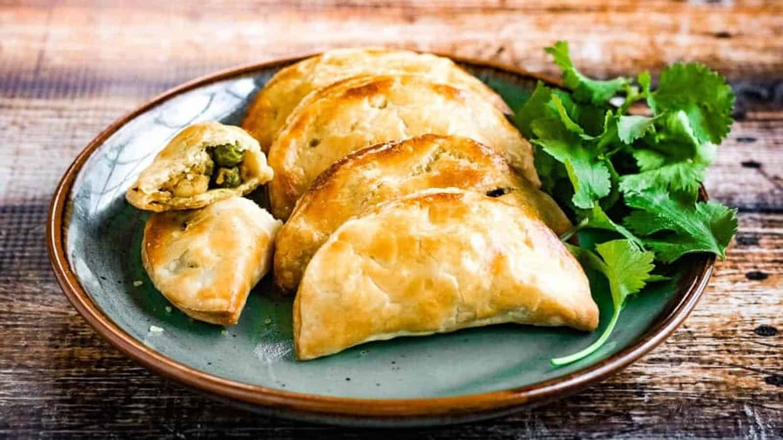 A plate of baked empanadas with a cilantro garnish, one empanada cut open to show a vegetable filling, on a rustic wooden table.