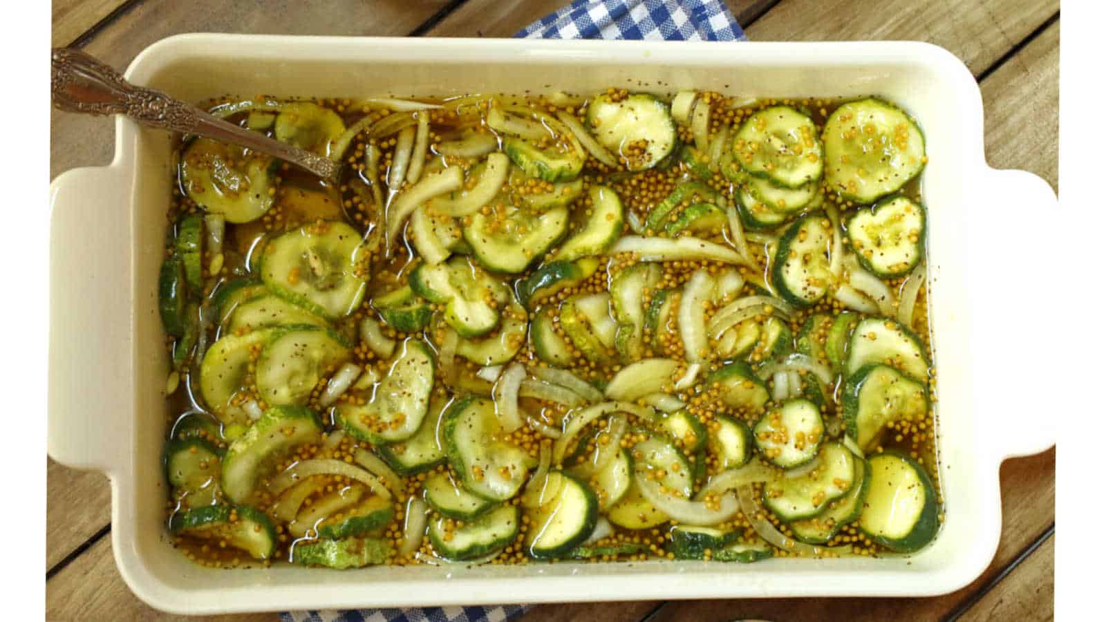 A white dish filled with sliced cucumbers, onions, and mustard seeds in a yellowish brine, with a spoon on one side.