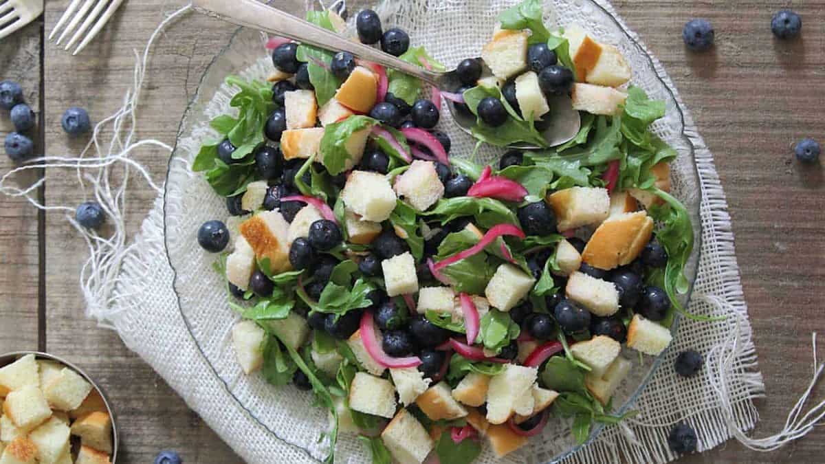 A plate of salad with arugula, blueberries, pickled onions, and bread cubes on a glass plate, set on a rustic wooden table.