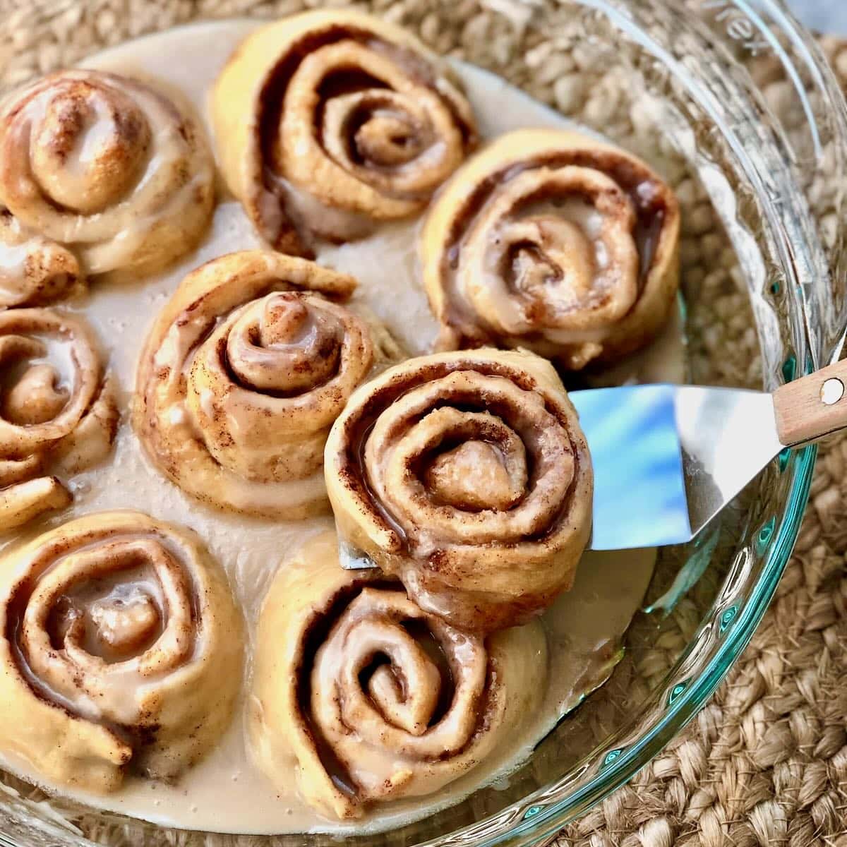 A glass dish filled with frosted cinnamon rolls, with one roll being lifted out using a metal spatula.