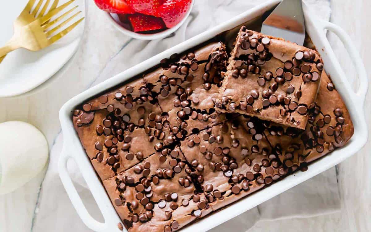 A rectangular baking dish filled with chocolate chip brownies, one piece slightly lifted, with a bowl of strawberries and a fork nearby.