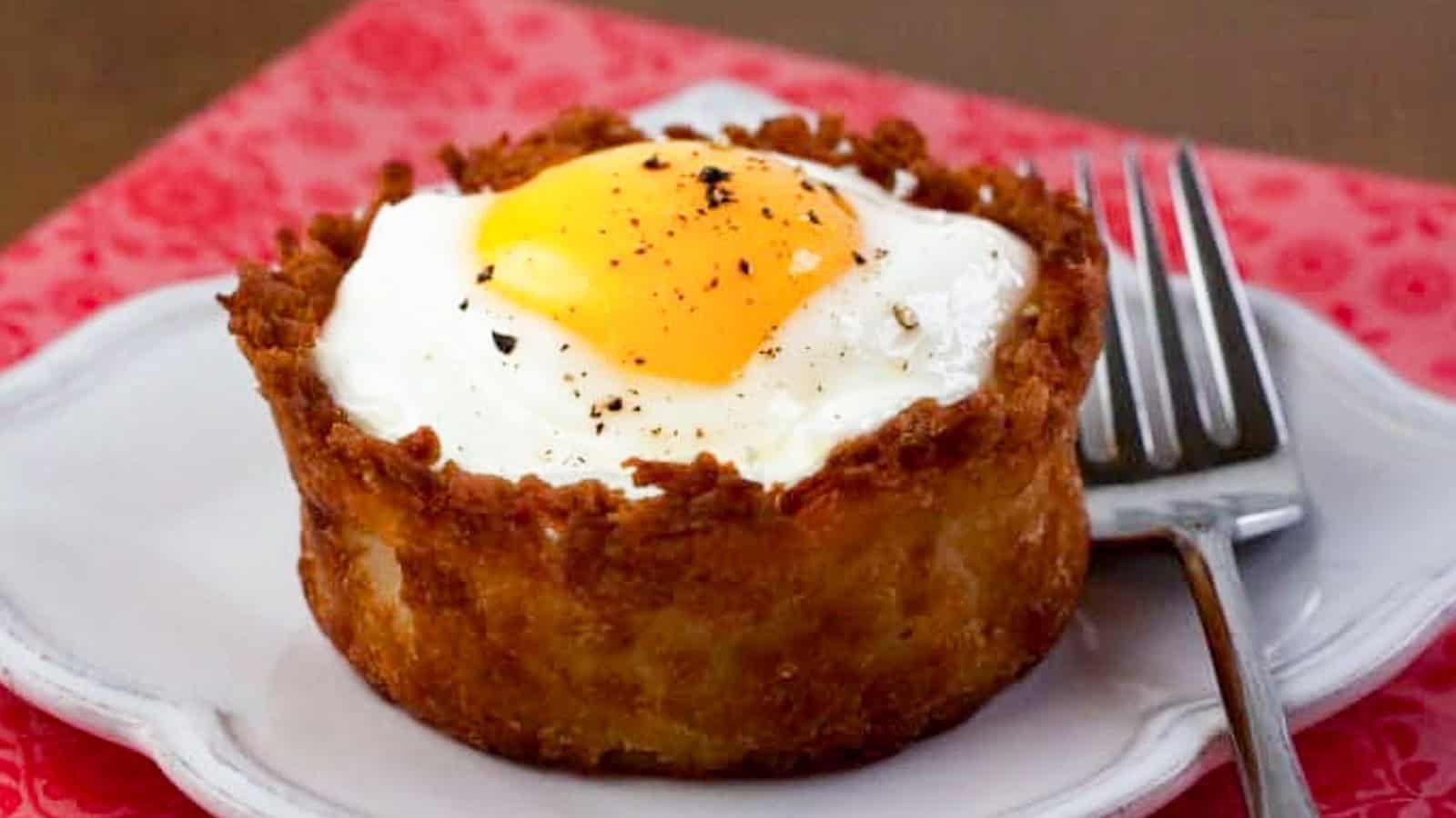 A baked breakfast cup with a crispy potato crust, topped with a sunny-side-up egg and black pepper, sits on a white plate next to a metal fork.