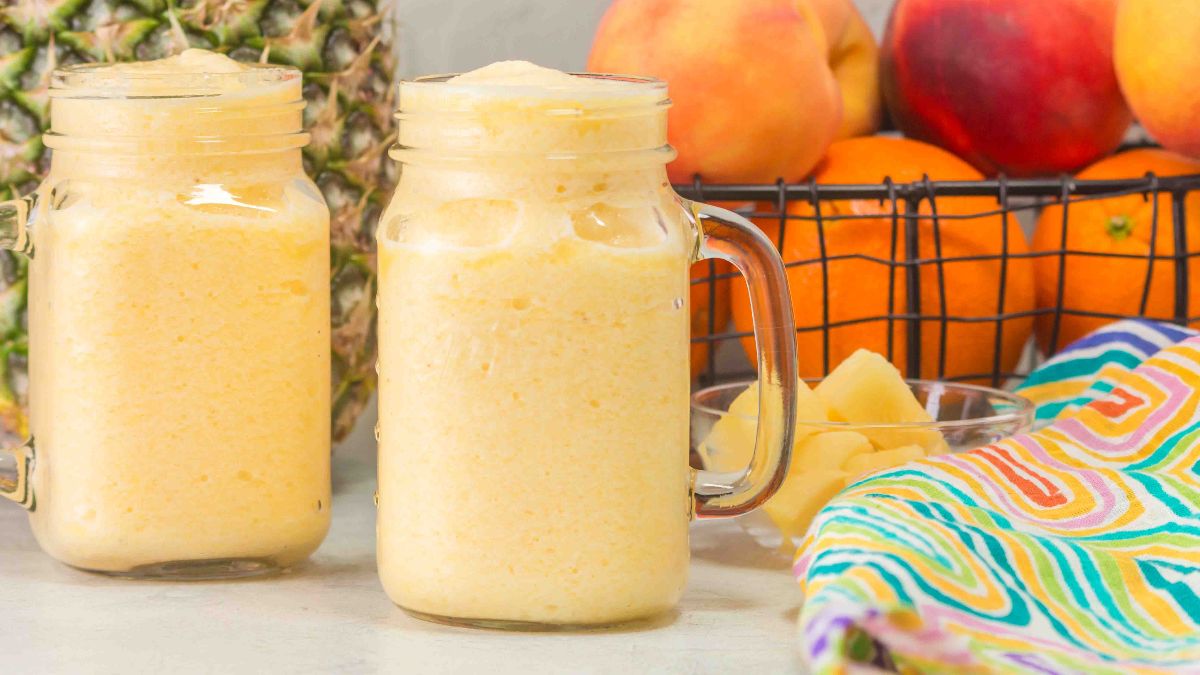 Two mason jars filled with a yellow smoothie are on a table next to a pineapple, a bowl of pineapple chunks, oranges, peaches, and a colorful cloth.