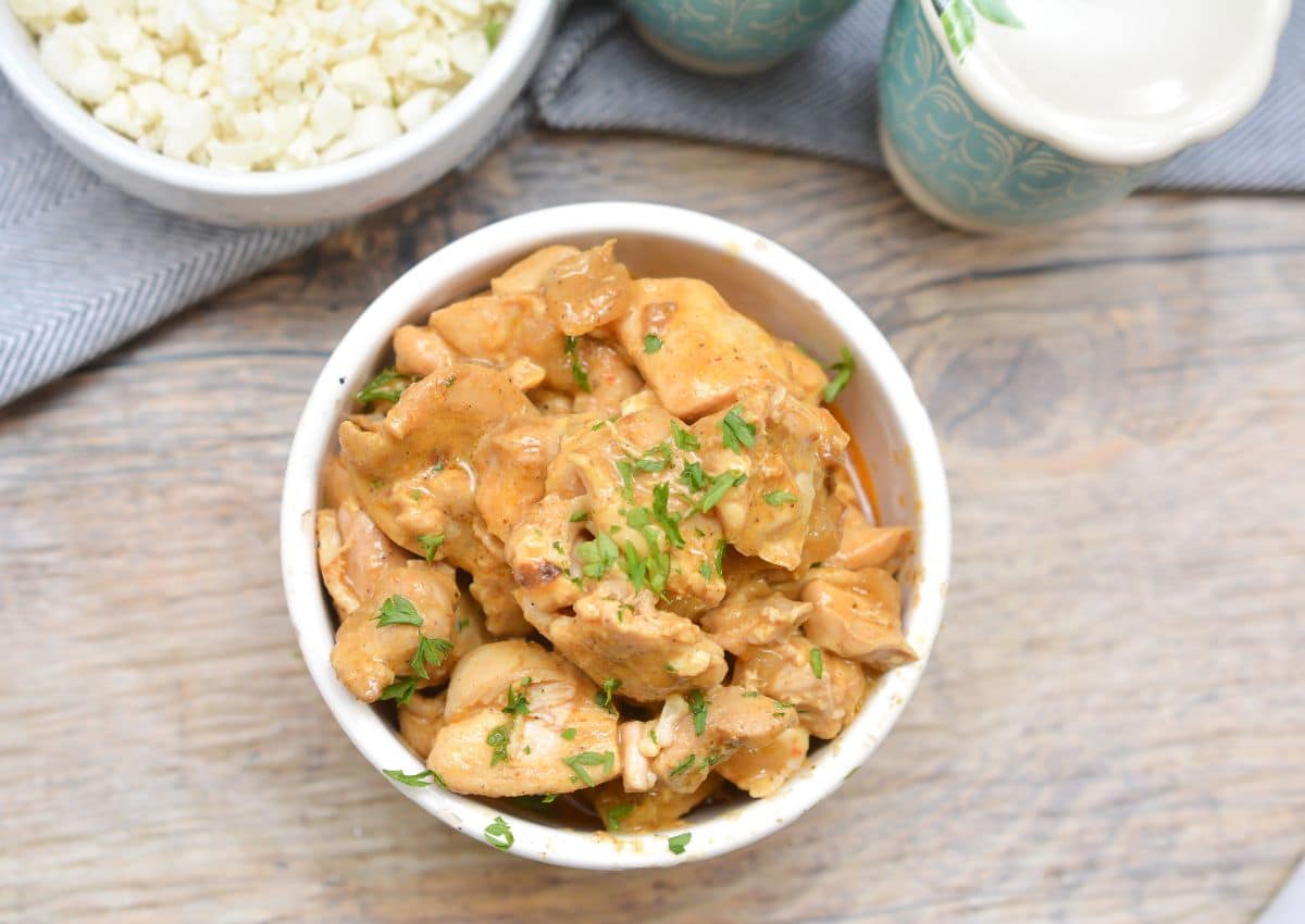 A bowl of cooked chicken pieces in a creamy sauce, garnished with chopped herbs, placed on a wooden surface near a bowl of cauliflower rice.
