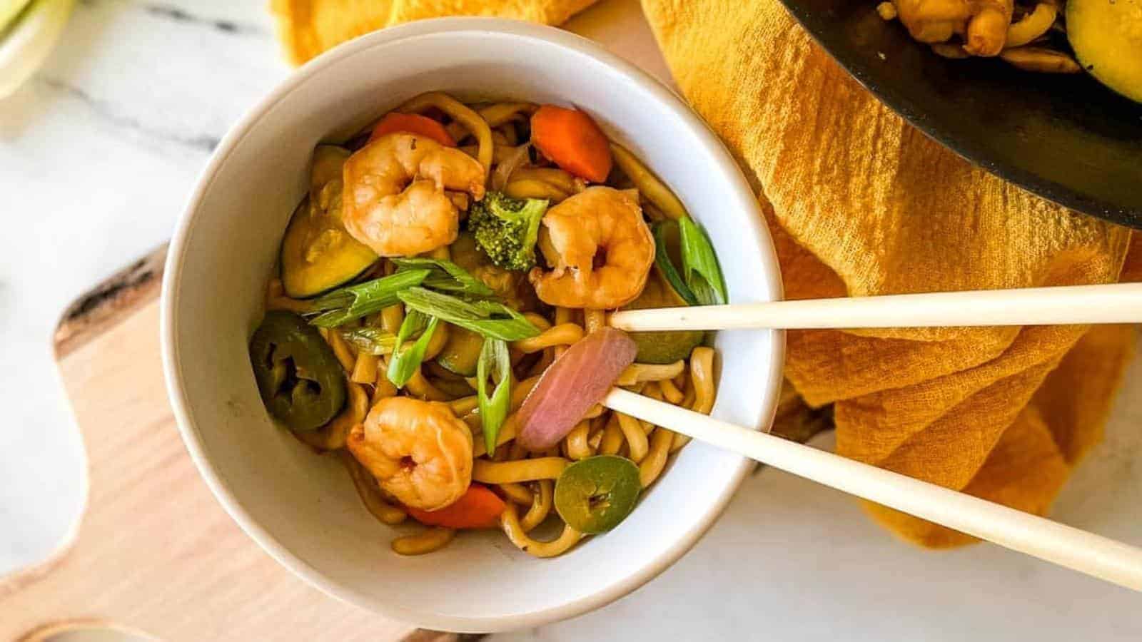 A bowl of shrimp stir-fry noodles with vegetables, garnished with green onions, and a pair of chopsticks resting on top.