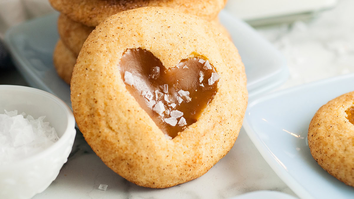 A round cookie with a heart-shaped cutout filled with caramel and topped with flaky sea salt, placed on a white surface next to other cookies and a small bowl of salt.