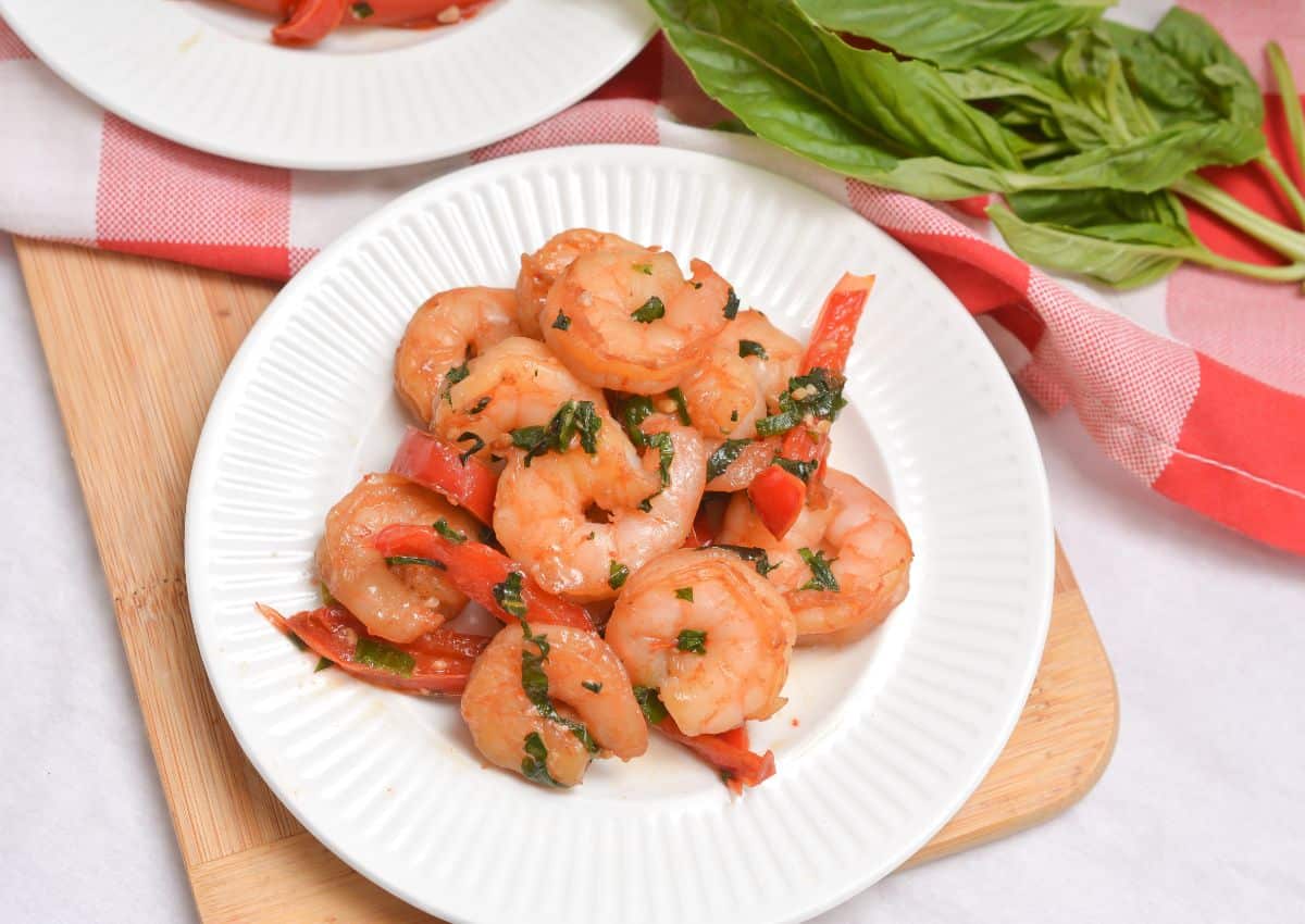 A white plate of cooked shrimp with sliced red peppers and fresh basil, placed on a wooden board next to a bunch of basil and a red-and-white checked cloth.