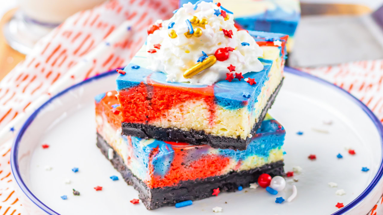 Two cheesecake bars with red, white, and blue marbled layers and chocolate crust, topped with whipped cream and festive sprinkles, on a white plate.