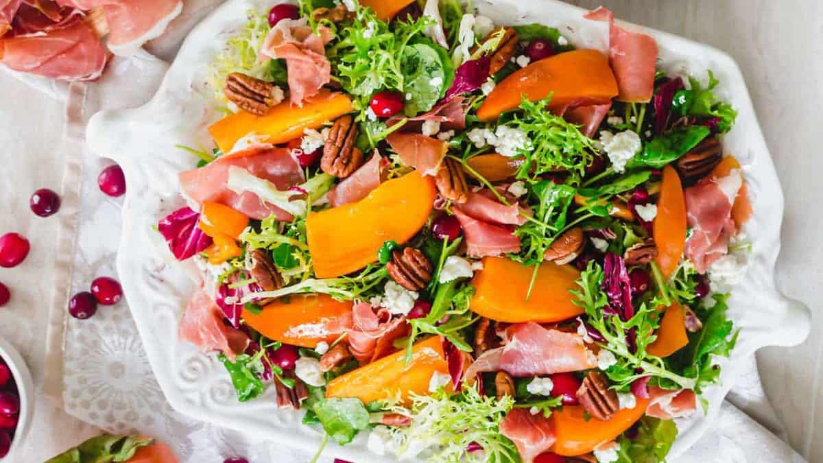 A salad on a white platter with mixed greens, persimmon slices, prosciutto, pecans, cranberries, and crumbled cheese.