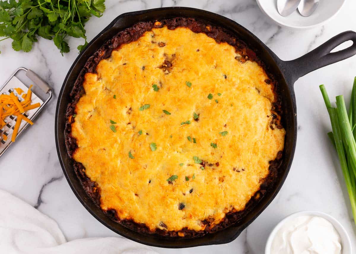 A cast iron skillet filled with cheesy cornbread-topped casserole, garnished with herbs, surrounded by green onions, grated cheese, cilantro, and a bowl of sour cream.