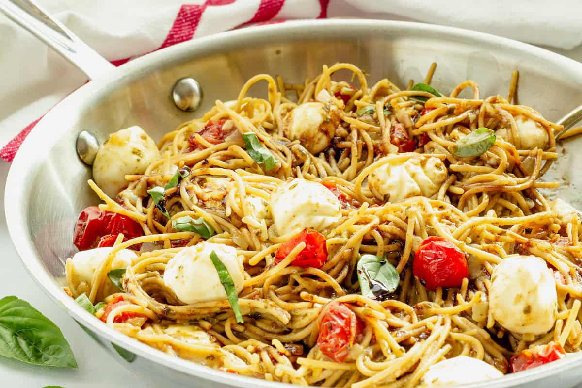 A stainless steel pan filled with spaghetti, cherry tomatoes, mozzarella balls, fresh basil, and a drizzle of balsamic glaze.