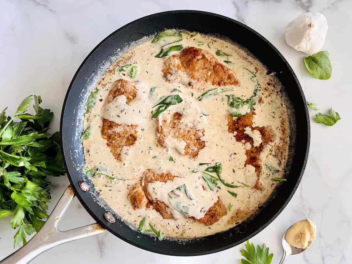 Chicken cutlets in a creamy sauce with spinach cooking in a black skillet, surrounded by fresh parsley, garlic, and a spoon with mustard on a marble surface.
