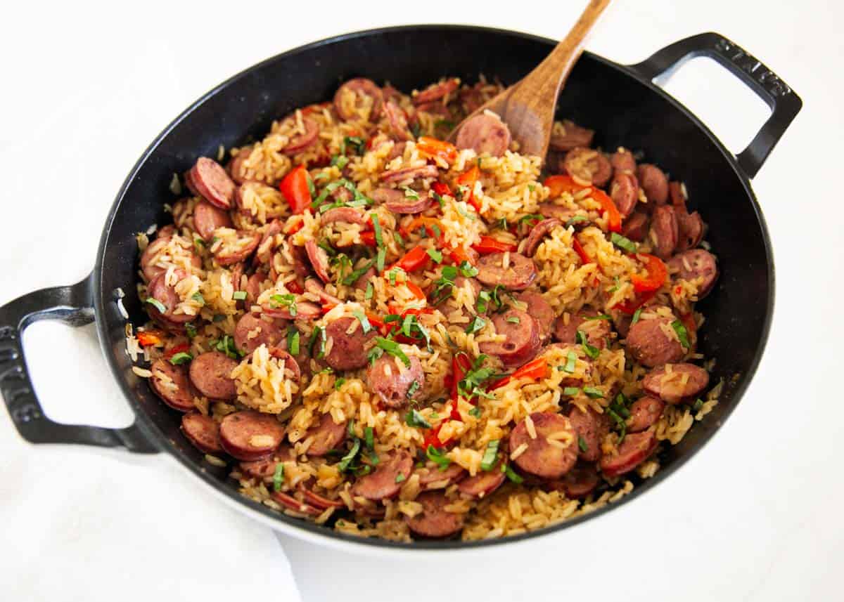 A skillet filled with rice, sliced sausage, red bell pepper, and herbs, with a wooden spoon resting inside.