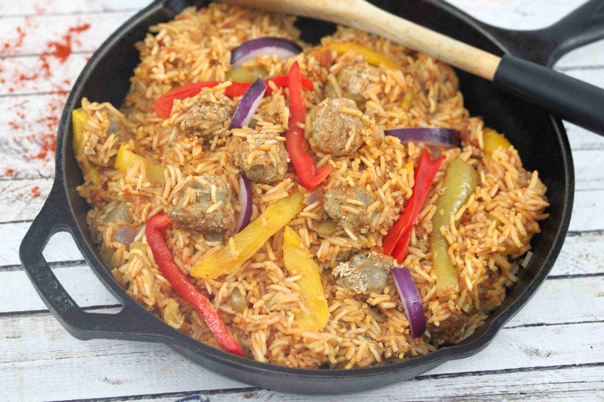 A cast iron skillet filled with cooked rice, sliced red and yellow bell peppers, red onion, and meatballs sits on a wooden surface with a wooden spoon resting on the side.