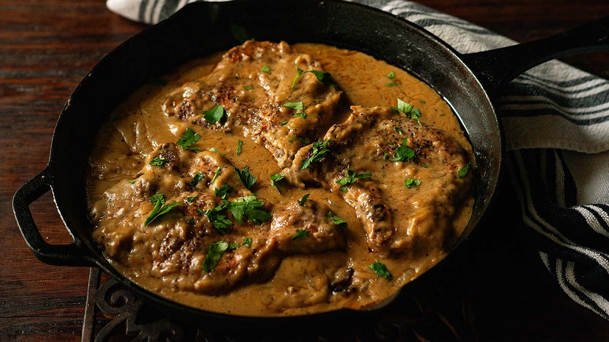 Pan-seared pork chops in a creamy brown sauce, garnished with chopped parsley, served in a cast iron skillet on a dark wooden table.