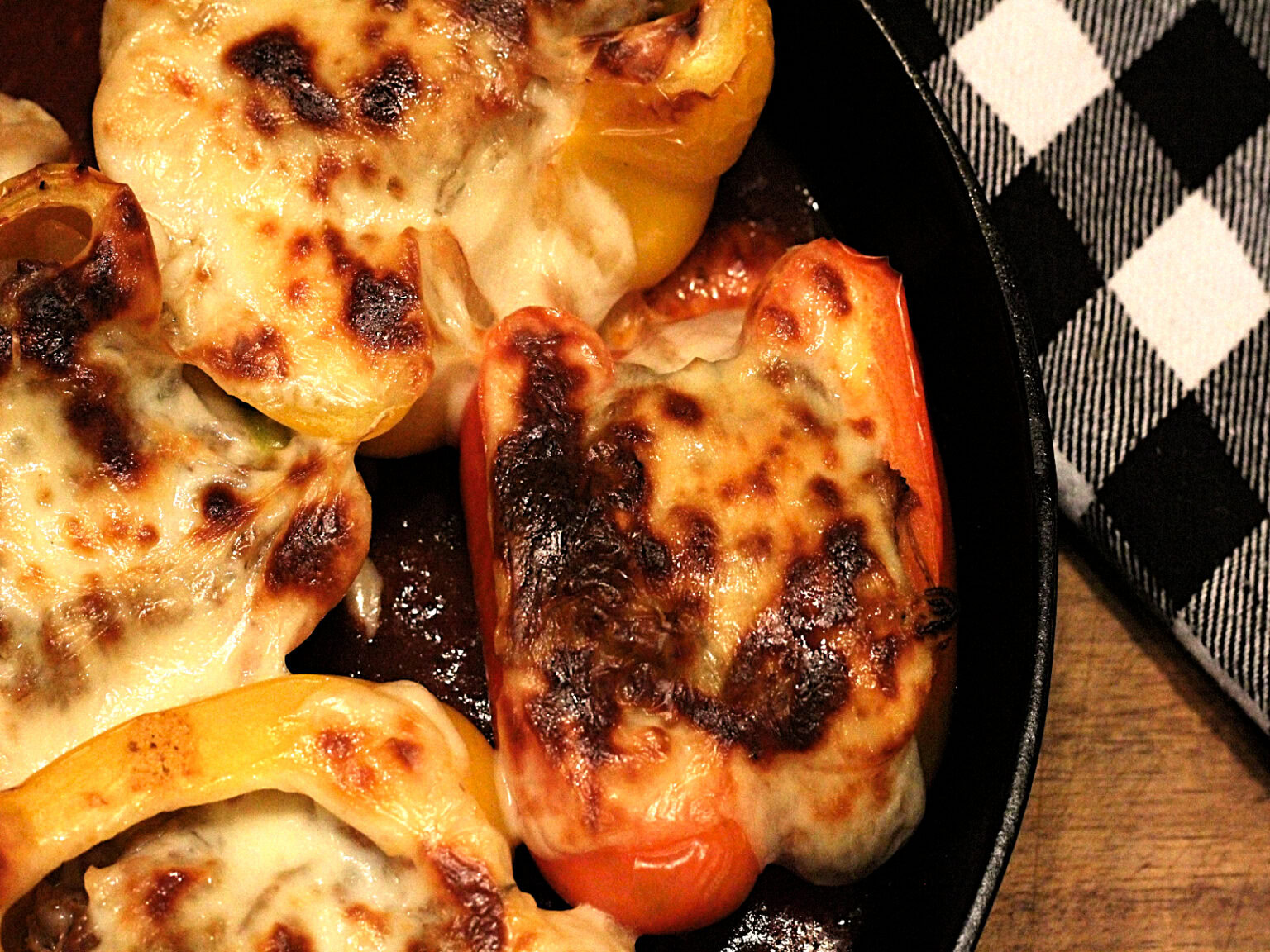 Stuffed bell peppers topped with melted, browned cheese in a skillet next to a black and white checkered napkin.
