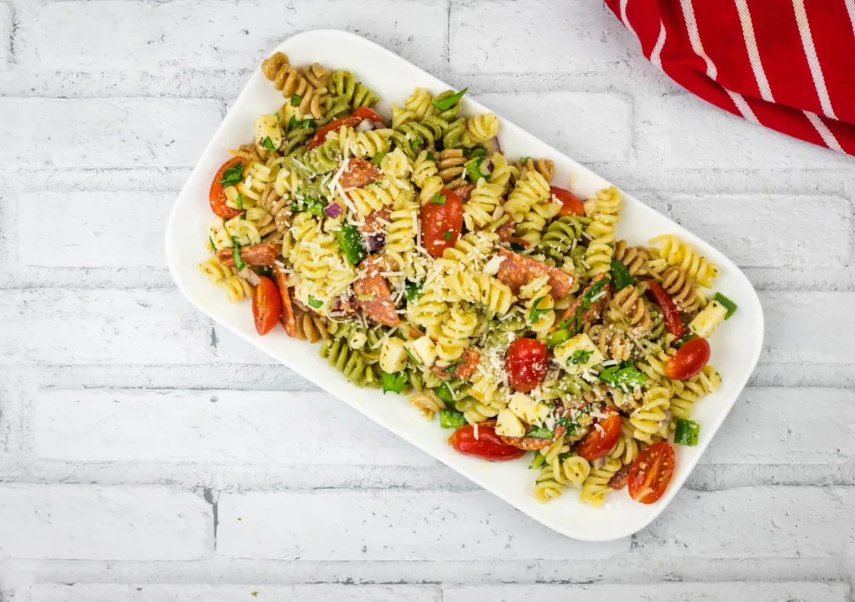 A rectangular white plate holds tri-color rotini pasta salad with cherry tomatoes, cheese cubes, greens, and shredded cheese on a white brick surface. A red striped cloth is nearby.