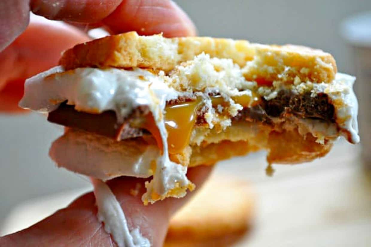 A hand holding a partially eaten smore with melted chocolate, caramel, and marshmallow between two cookies.