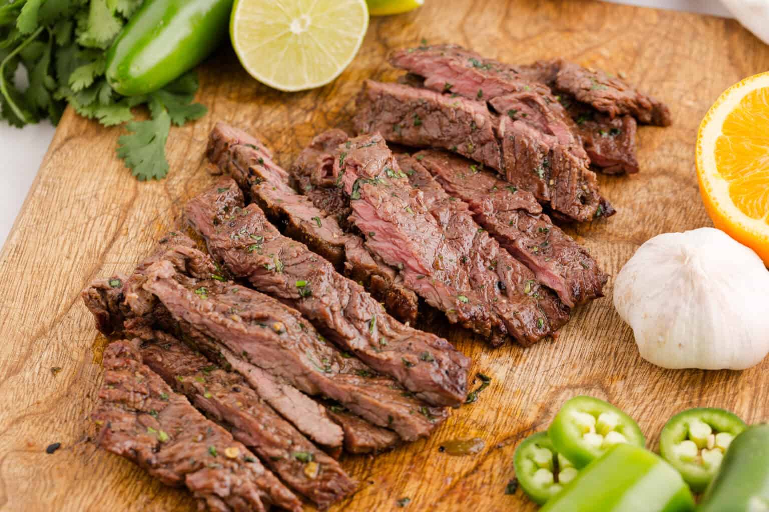 Sliced grilled steak on a wooden cutting board, surrounded by lime, orange, garlic, cilantro, and sliced jalapeรฑos.