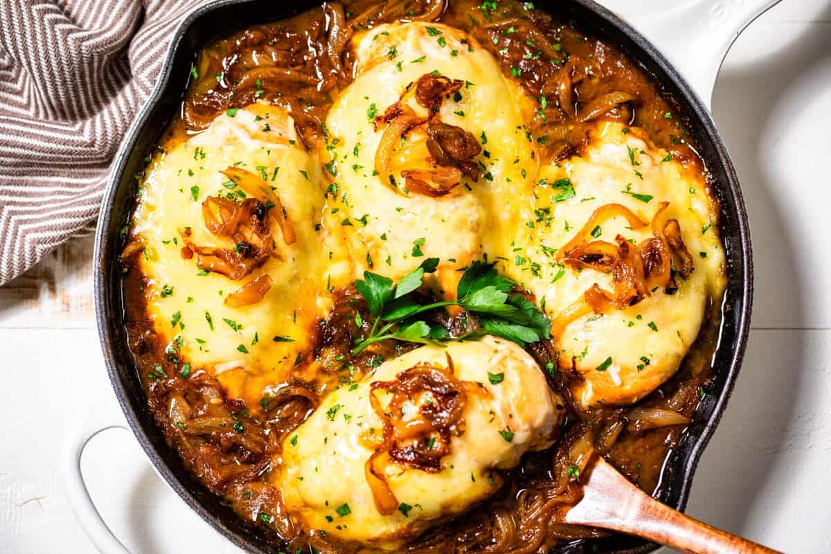 Four pieces of chicken breast topped with melted cheese and caramelized onions in a skillet with a brown sauce, garnished with chopped parsley.