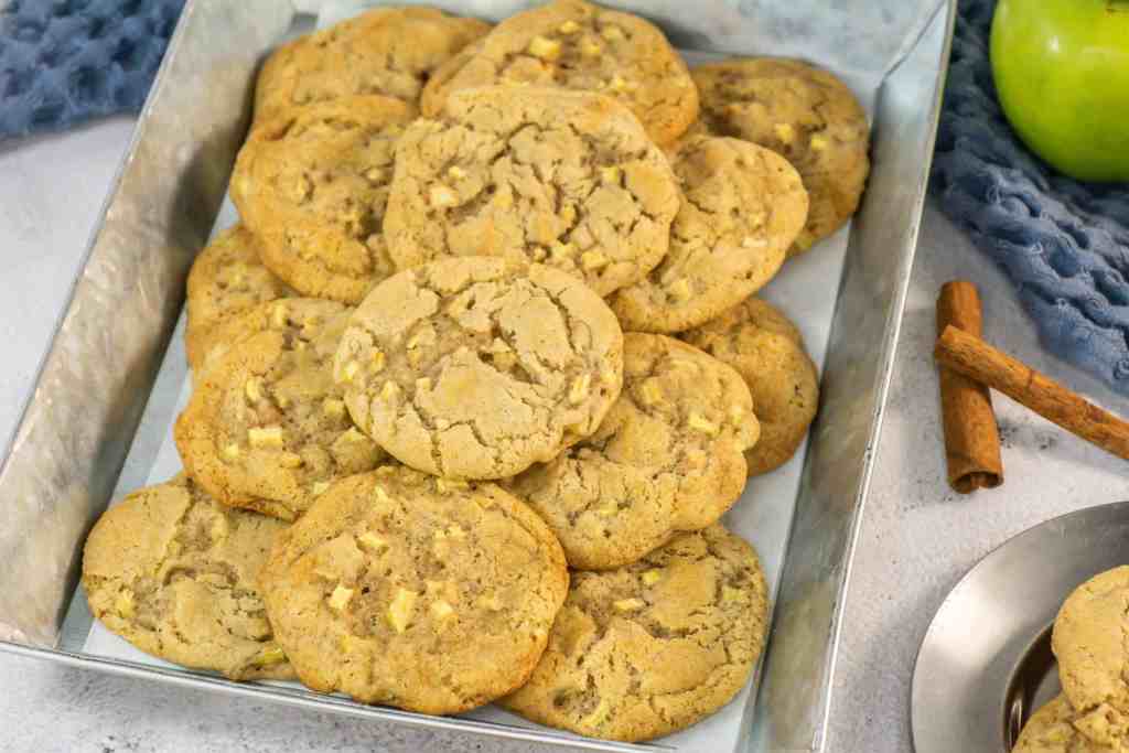 A tray filled with a stack of homemade cookies containing visible apple chunks sits on a countertop next to cinnamon sticks and a green apple.