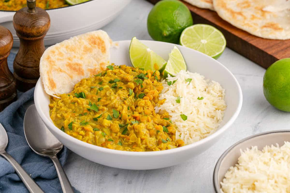 A white bowl filled with yellow lentil curry, white rice, lime wedges, and a piece of flatbread, with extra rice and limes in the background.