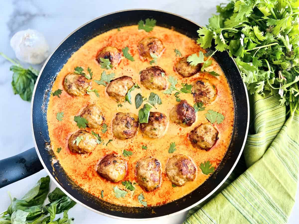 A skillet filled with meatballs in a creamy orange sauce, garnished with fresh cilantro, sits next to a bunch of herbs and a green striped towel.