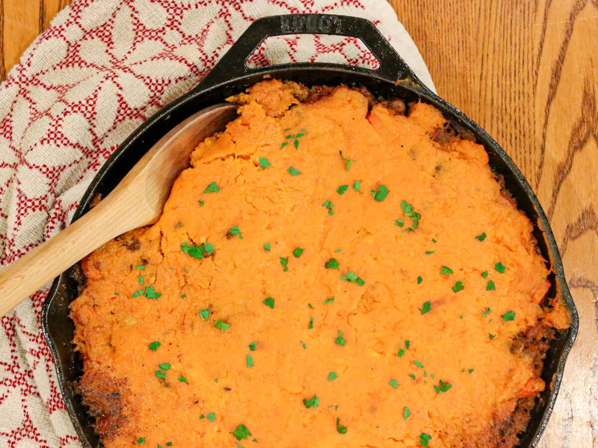 A cast iron skillet filled with a baked sweet potato casserole, garnished with chopped herbs, sits on a wooden table next to a patterned cloth and a wooden spoon.