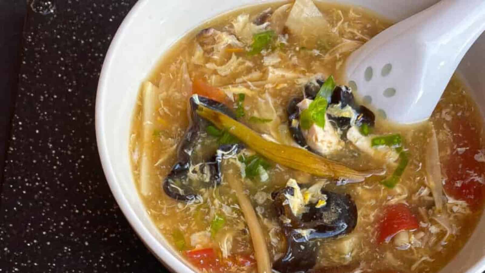 A bowl of hot and sour soup with mushrooms, shredded vegetables, egg ribbons, and a white soup spoon on the side.