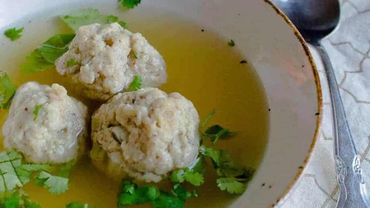 Three matzo balls in clear broth, garnished with fresh cilantro, served in a shallow white bowl with a spoon beside it.