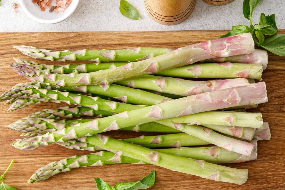 Fresh green asparagus spears are arranged on a wooden cutting board, with basil leaves, a bowl of pink salt, and pepper mills nearby.