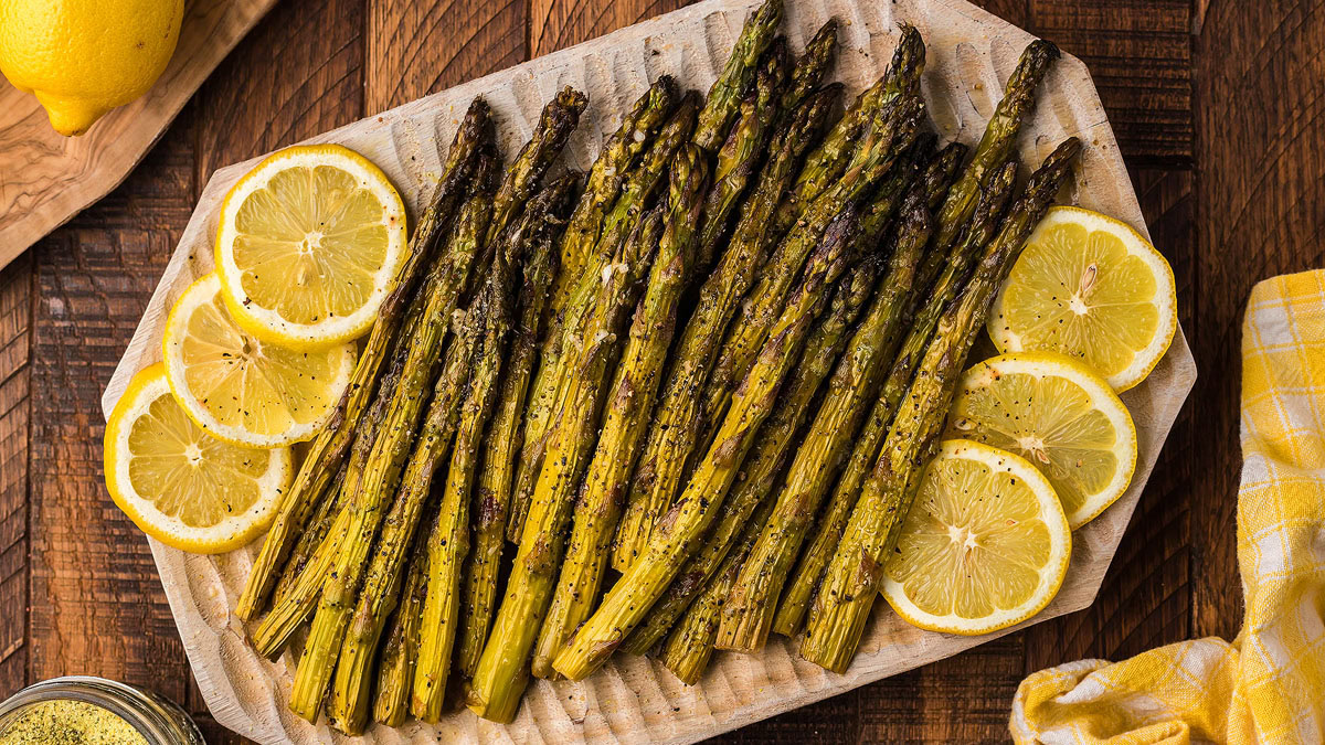 Grilled asparagus spears arranged on a wooden platter with slices of lemon, placed on a wooden table.