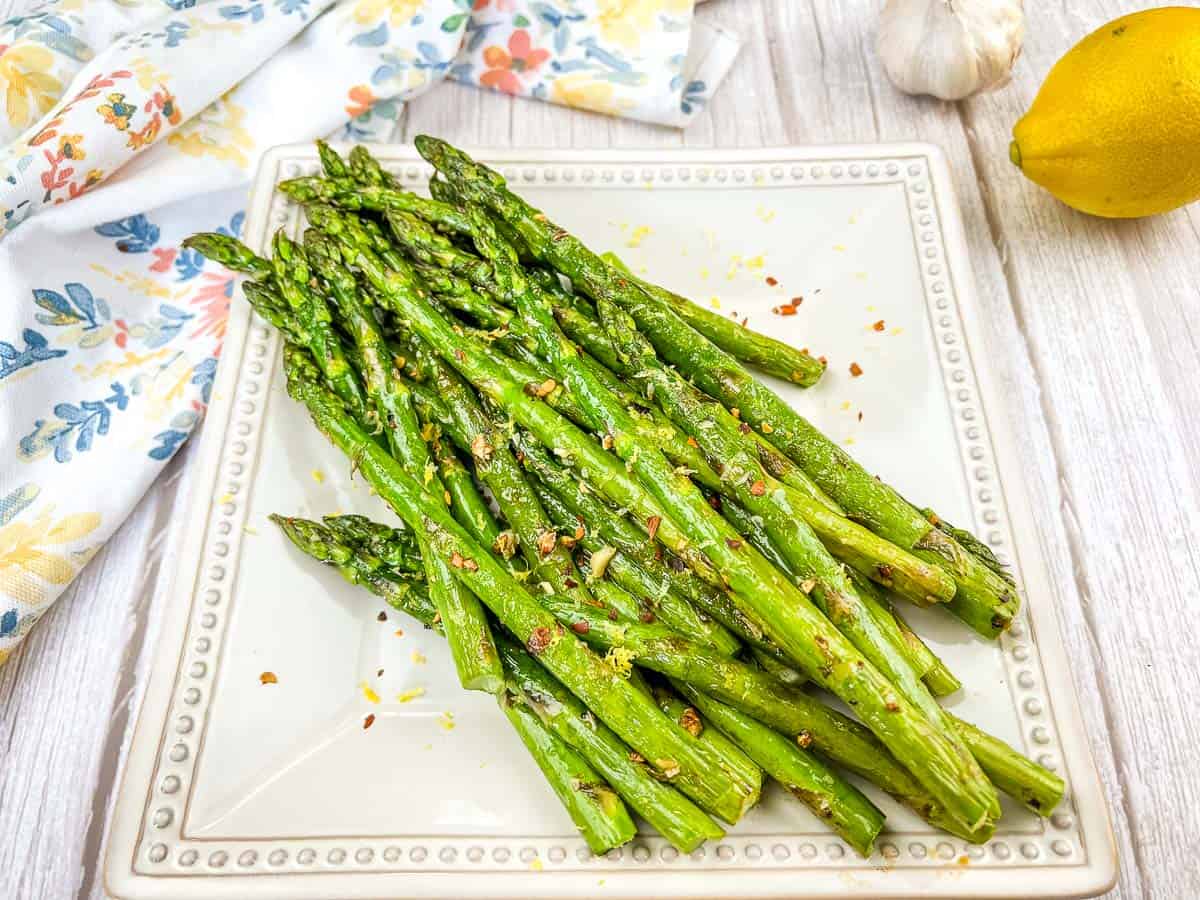 A square plate of cooked asparagus spears seasoned with spices, set on a light wooden surface with a lemon, garlic, and a floral napkin nearby.