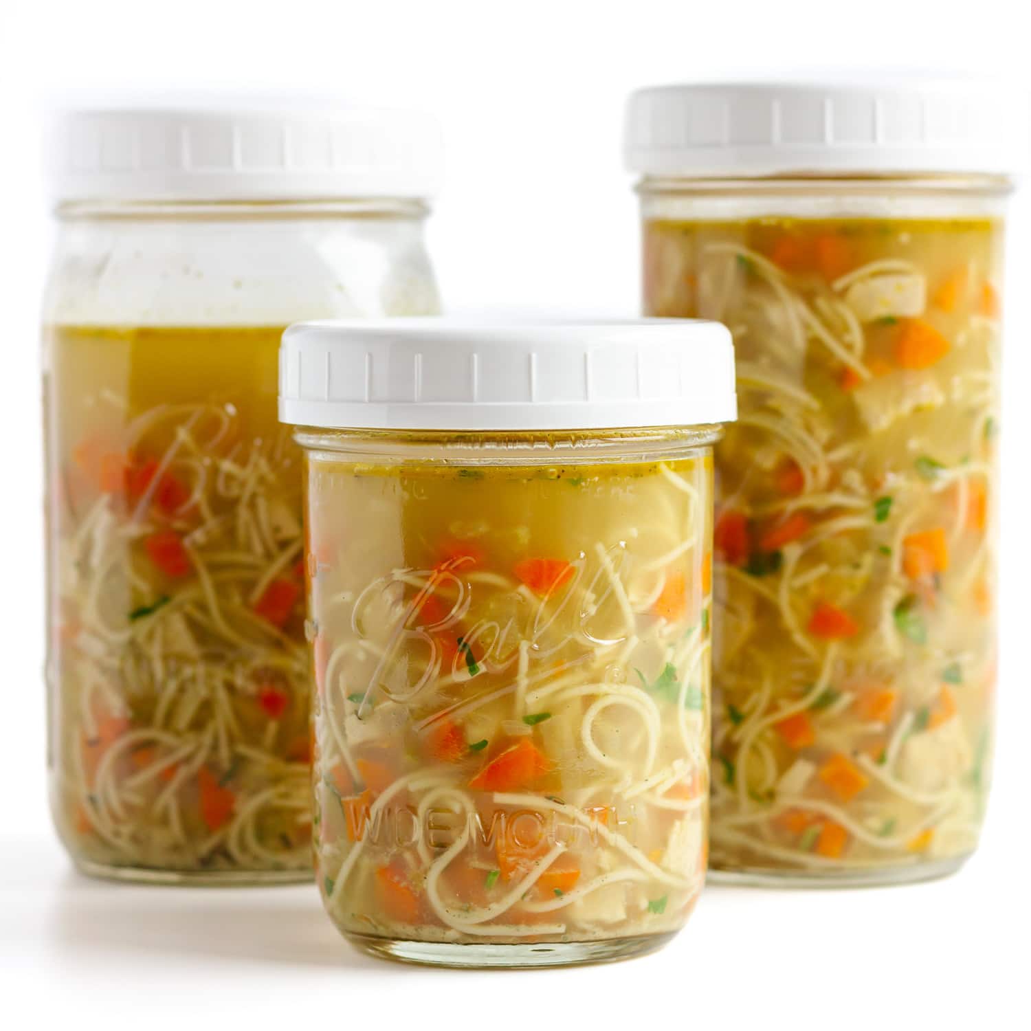 Three glass jars with white lids are filled with homemade chicken noodle soup, featuring visible noodles, carrots, and broth.