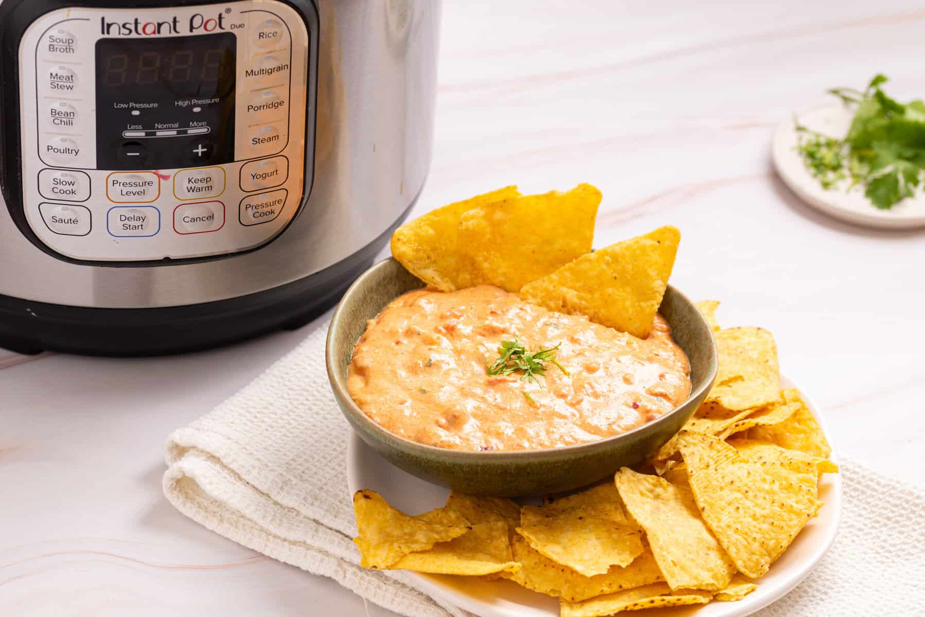 A bowl of cheese queso dip garnished with herbs and tortilla chips, placed next to an Instant Pot on a white surface.