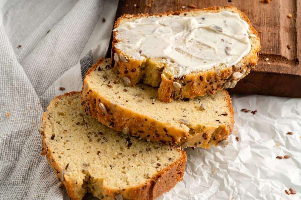 Three slices of seeded bread are on parchment paper, with one slice spread with a layer of cream cheese. A wooden board is in the background.