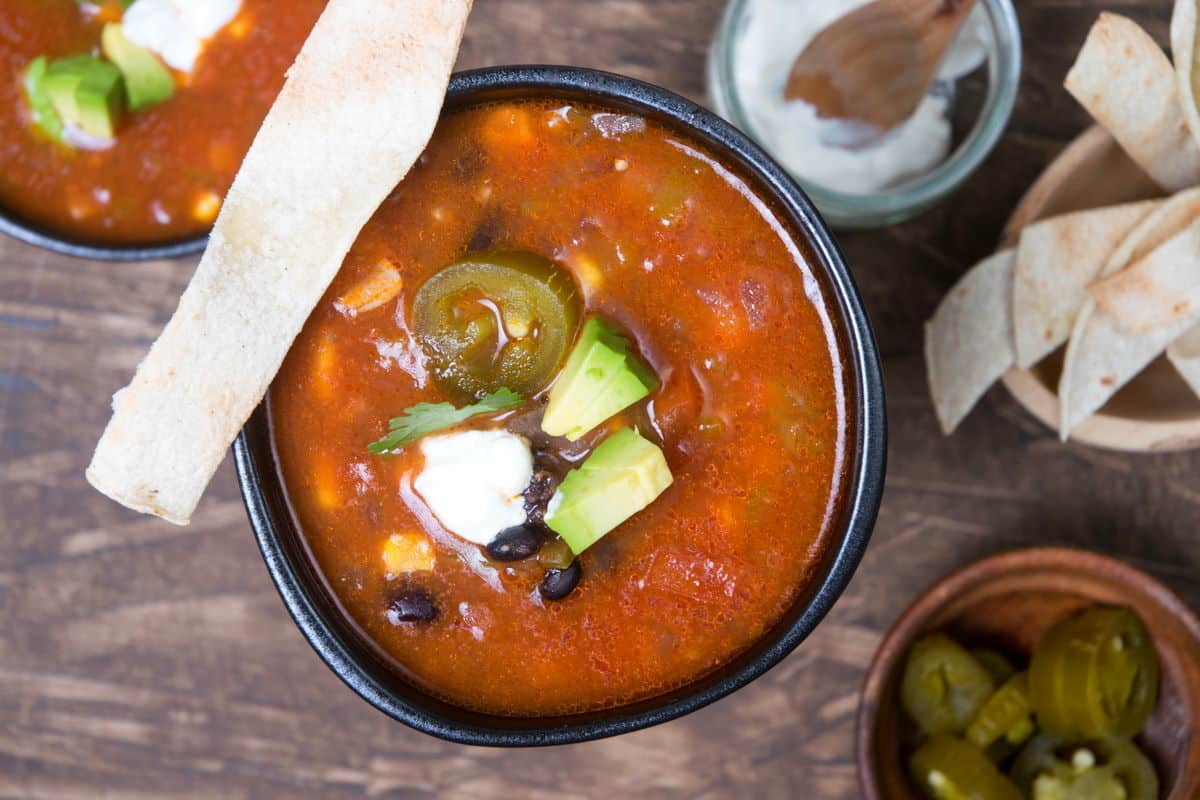A bowl of tomato-based soup topped with avocado, jalapeรฑo slices, sour cream, and cilantro, served with a tortilla strip, alongside small bowls of tortilla strips and jalapeรฑos.