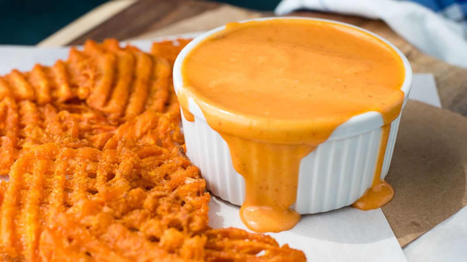 Waffle-cut sweet potato fries served with a ramekin of orange dipping sauce overflowing onto the surface.