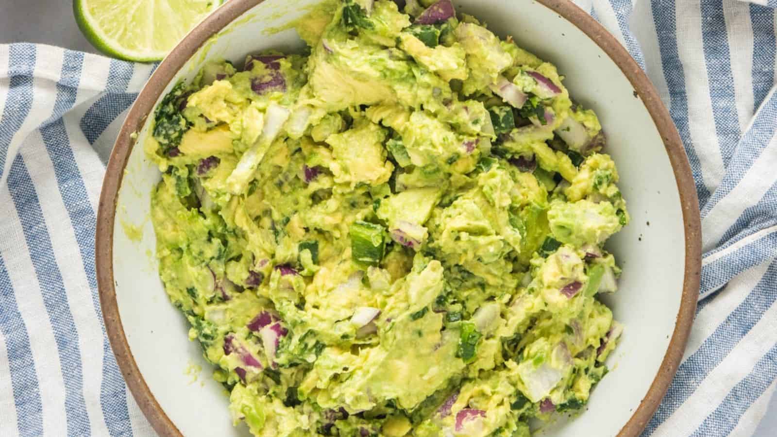A bowl of chunky guacamole with chopped onions, cilantro, and avocado, placed on a striped cloth with a lime wedge nearby.