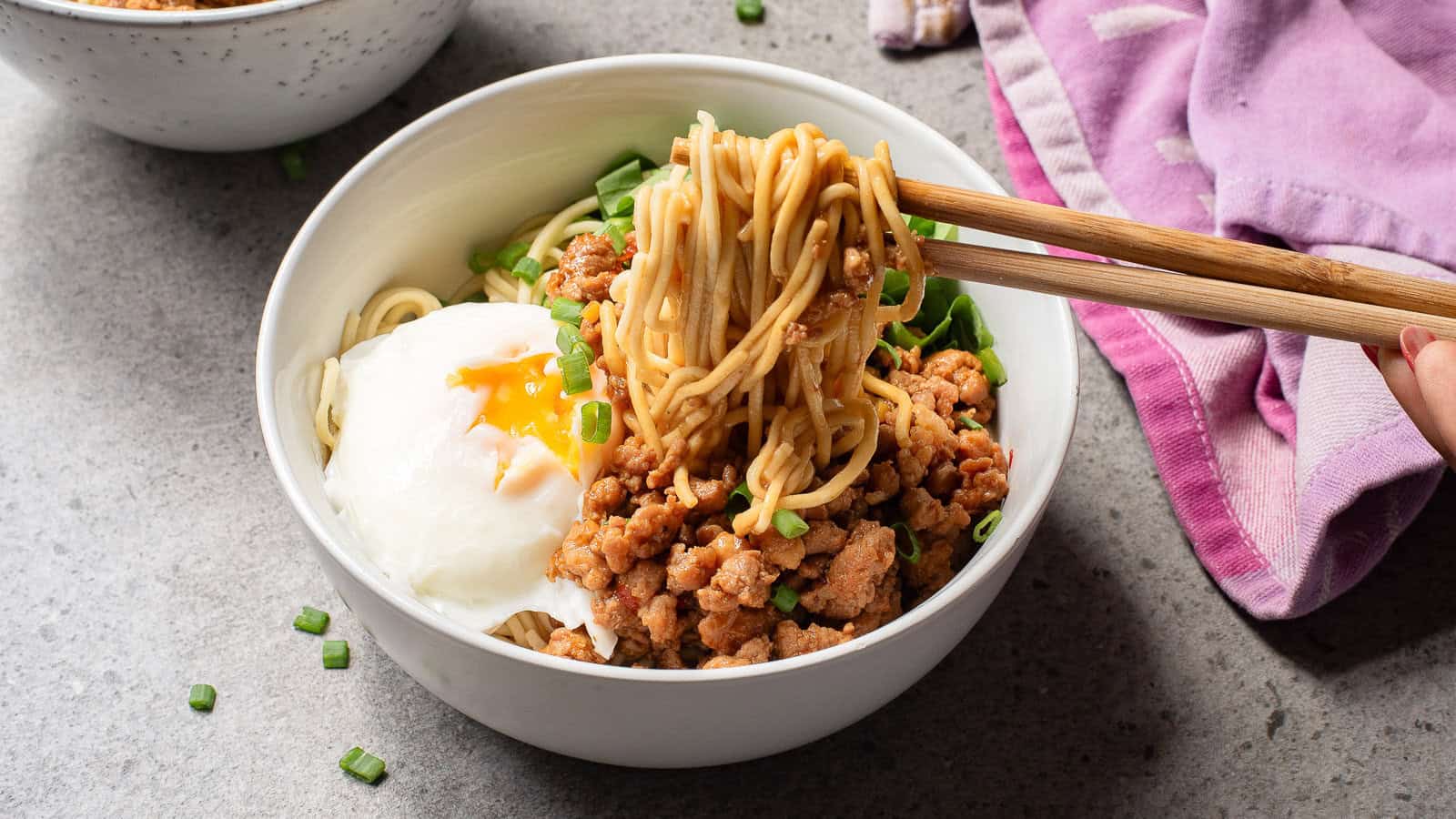 A bowl of noodles topped with ground meat, chopped green onions, and a soft-boiled egg; chopsticks lift noodles from the bowl.