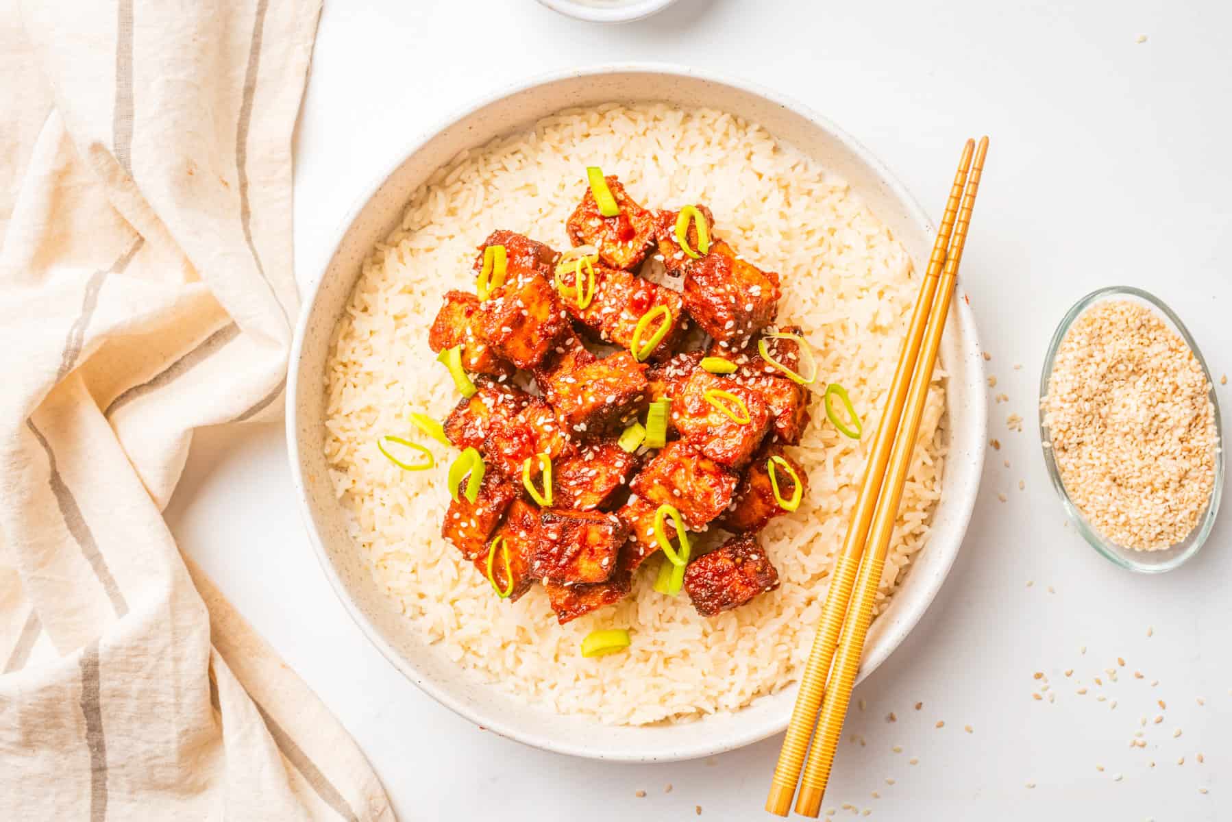 A bowl of white rice topped with glazed tofu cubes, chopped green onions, and sesame seeds, with chopsticks resting on the side.