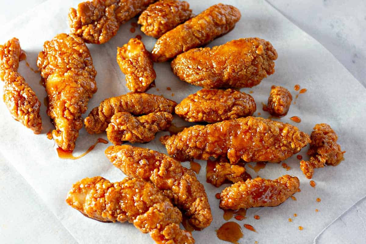 Crispy fried chicken tenders with a sticky glaze are arranged on white parchment paper, with sauce drizzled across the surface.