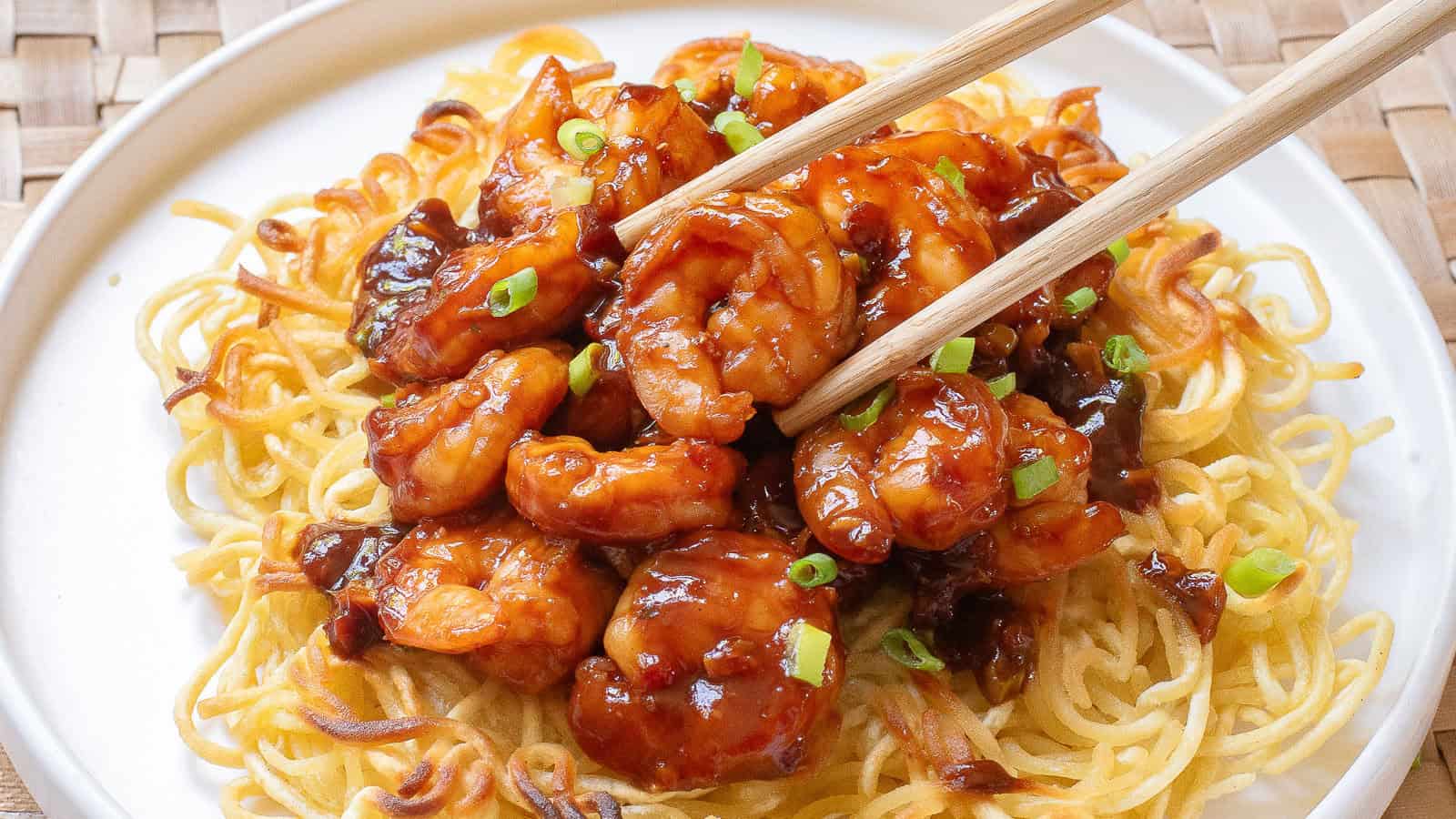 A plate of crispy noodles topped with glazed shrimp and chopped green onions, with chopsticks picking up a shrimp.