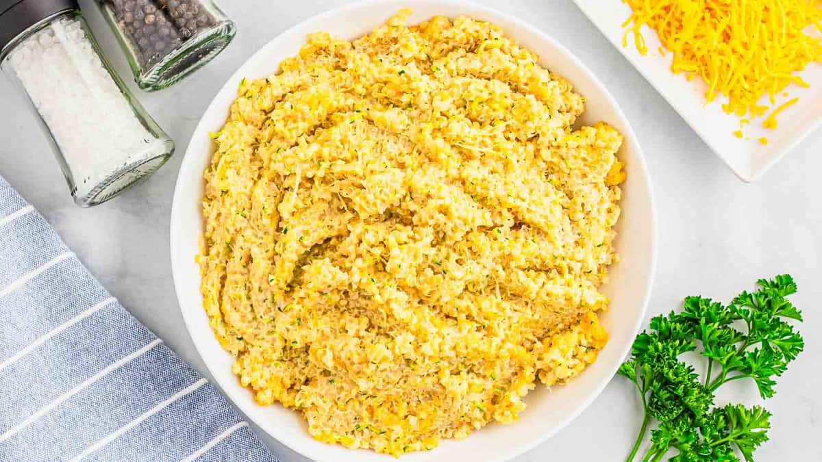 A bowl of cheesy, seasoned quinoa sits on a white surface next to a striped towel, fresh parsley, shredded cheese, and salt and pepper shakers.
