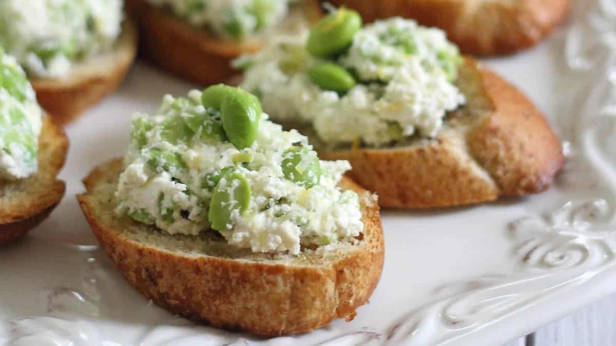 Slices of toasted bread topped with ricotta cheese and green edamame beans, arranged on a white plate.