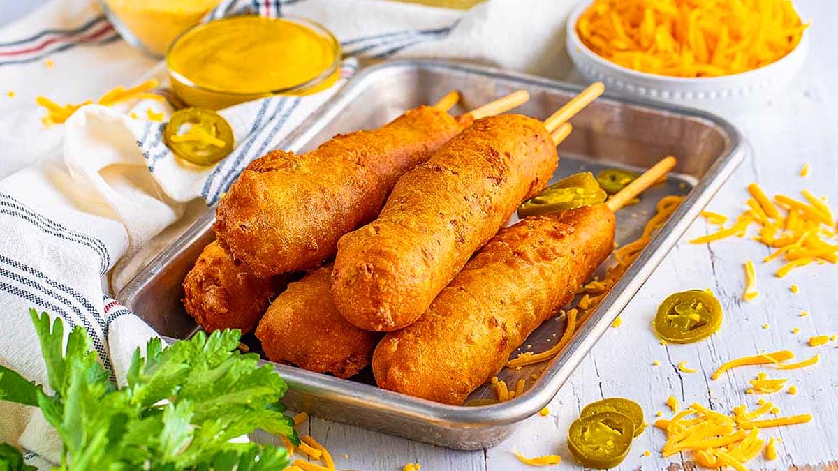 A metal tray with four corn dogs on sticks, surrounded by shredded cheese, jalapeรฑo slices, a bowl of mustard, and a bunch of parsley on a white wooden surface.