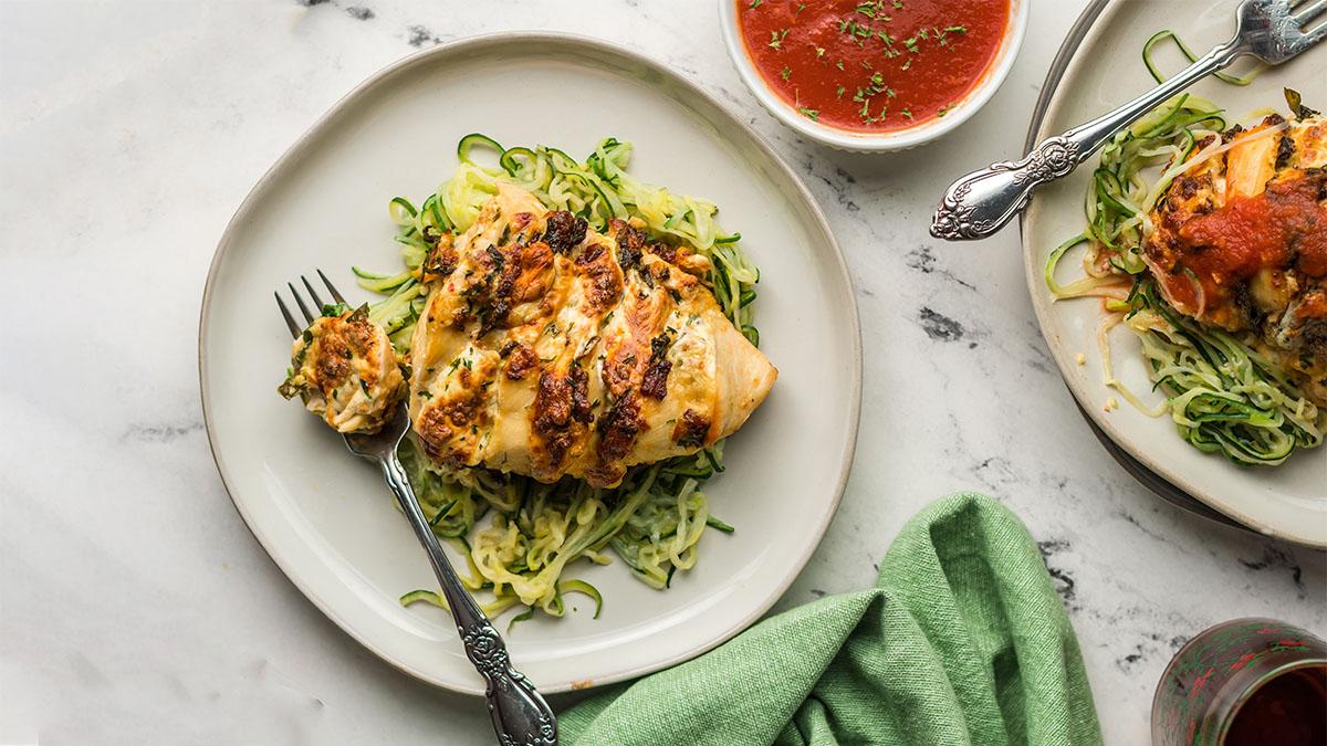 Grilled stuffed chicken breast served on a bed of spiralized zucchini noodles, with a forkful of chicken and a bowl of tomato sauce on the side.